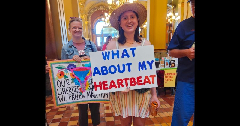 Photo is of signs held by two women at the protest of the 2023 near-total abortion ban. Both are in the Capitol Rotunda. The woman in the center is wearing a hat and smiling over her sign that reads "what about my heartbeat." On the left, just behind her, is a woman in a jean jacket who is also smiling and holding a sign that reads "Our liberties we prize and our rights we will maintain." The words are around an illustration of a uterus that's surrounded by flowers.