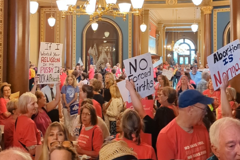 A packed crowd of people in the Captiol Rotunda in 2023, on the day the abortion ban passed in a special session, pink is the predominant color. Three signs are visible: one says 'abortion is normal,' one says 'no forced births' and one says 'if I wanted your religion in my pussy I'd fuck a priest.'