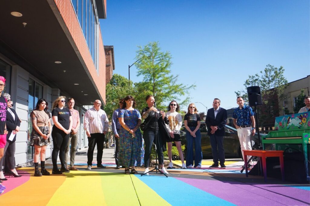 A picture from the official presentation of the Pride mural in the East Village, a line of people stand on the rainbow mural with artist Cat Rocketship at the mic.