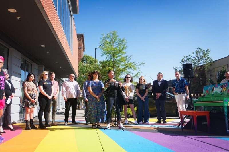 A picture from the official presentation of the Pride mural in the East Village, a line of people stand on the rainbow mural with artist Cat Rocketship at the mic.
