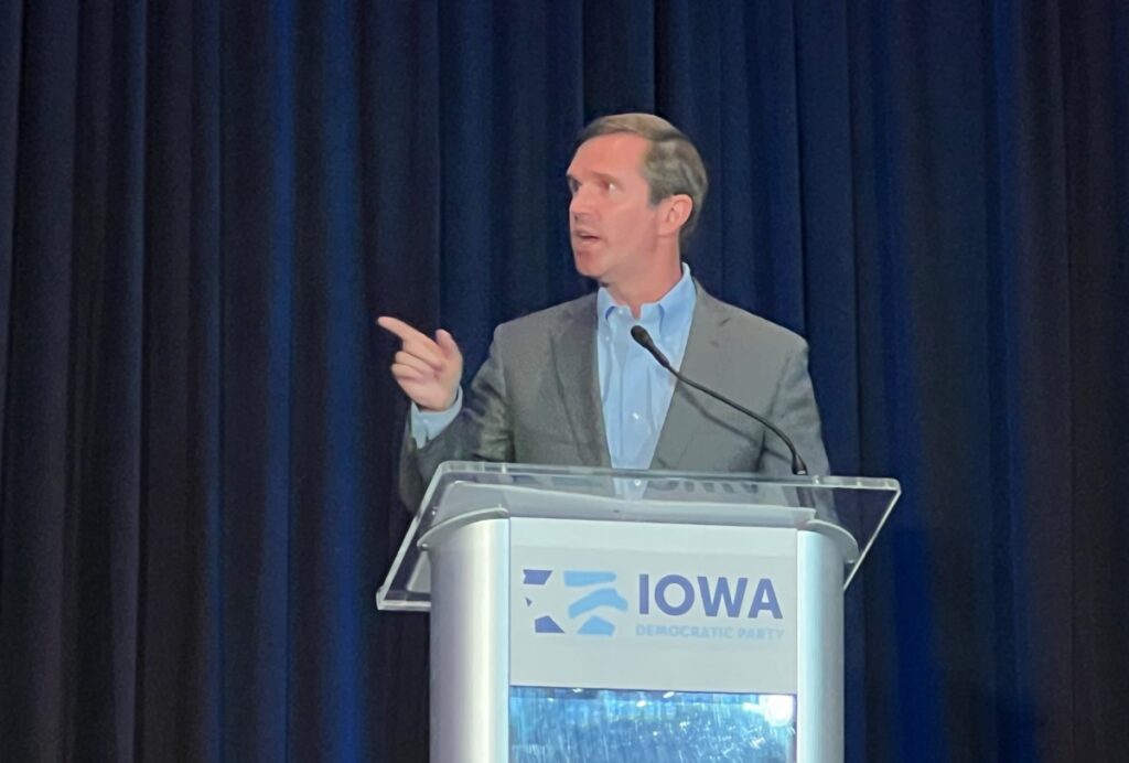 Gov. Andy Beshear at a clear podium that reads "Iowa Democratic Party" in blue. Beshear is pointing while he talks about the 2024 election.