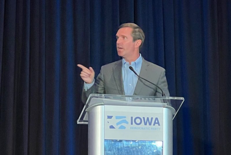 Gov. Andy Beshear at a clear podium that reads "Iowa Democratic Party" in blue. Beshear is pointing while he talks about the 2024 election.