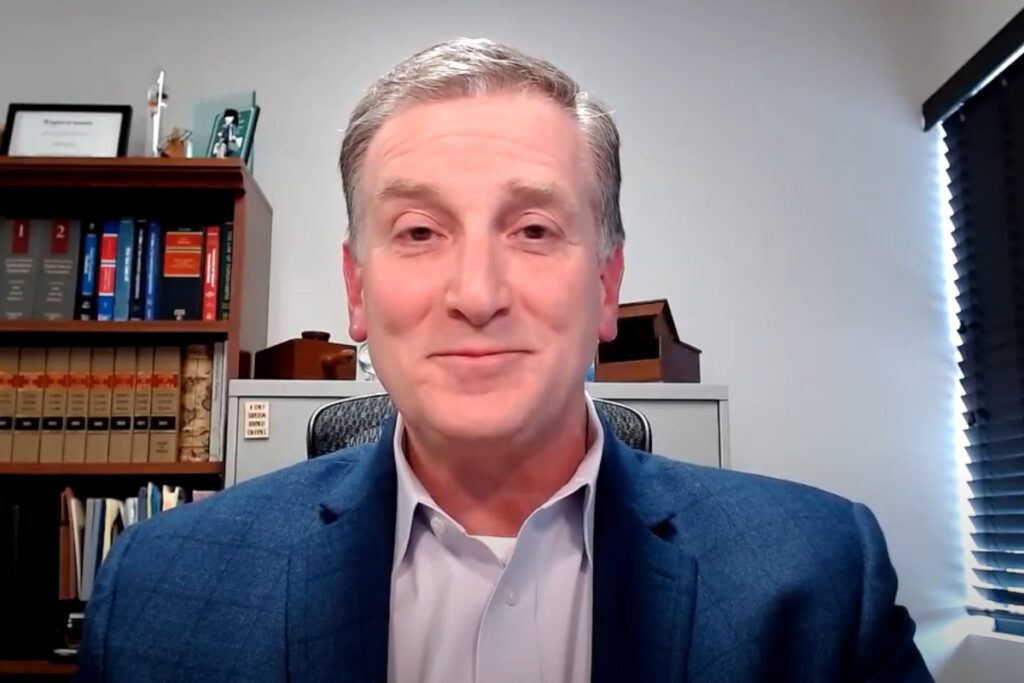 Bill Gustoff, a white man, is smiling with a closed mouth in an office. There's a bookshelf behind him.