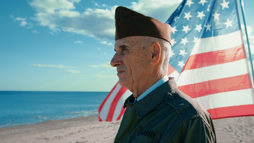 United States Veteran Looks At The Ocean From The Beach With America Flag behind him.