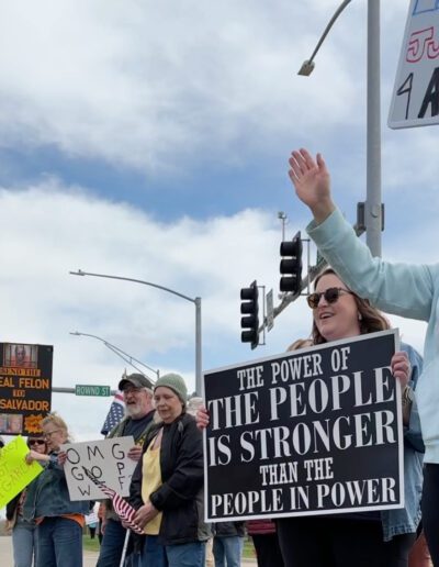 People wave signs protesting the Trump administration and DOGE and in support of democracy during a Day of Action rally along a stretch of Greenhill Road in Cedar Falls on Saturday, April 19, 2025. (Amie Rivers/Iowa Starting Line)