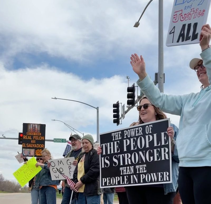 People wave signs protesting the Trump administration and DOGE and in support of democracy during a Day of Action rally along a stretch of Greenhill Road in Cedar Falls on Saturday, April 19, 2025. (Amie Rivers/Iowa Starting Line)