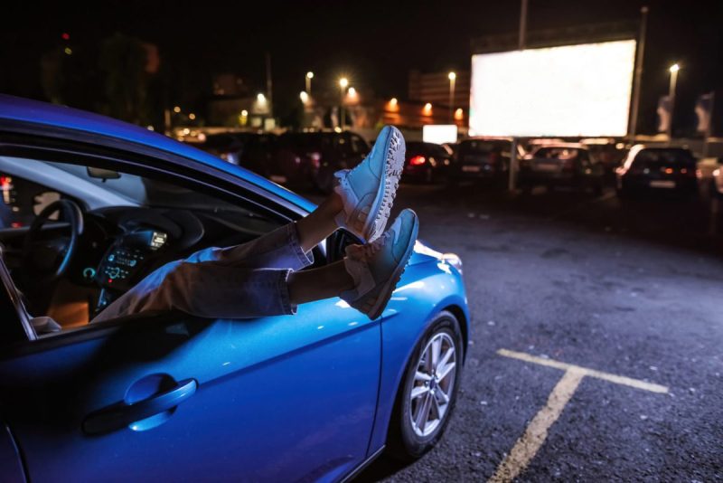 legs and feet dangle outside of an open passenger side window in front of a drive in movie theater screen