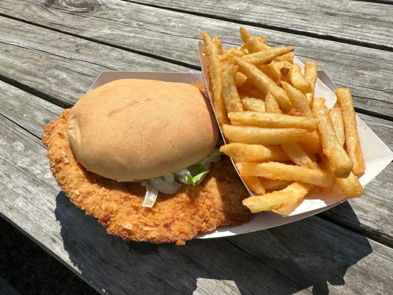 a fried pork tenderloin sandwich and french fries in a paper tray on a wooden table outside